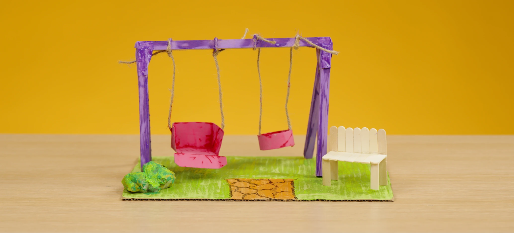 Swing structure made from lengths of wood with cardboard seats and a small bench made from lollipop sticks.