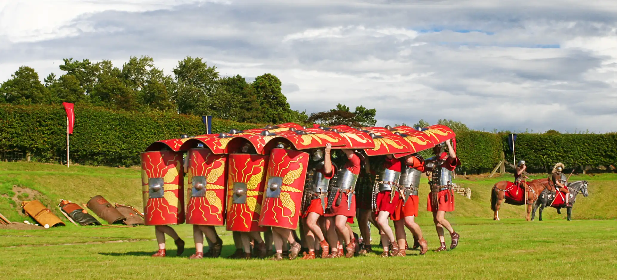 Roman soldiers in red shields demonstrate the testudo formation during a re-enactment, with cavalry in the background.