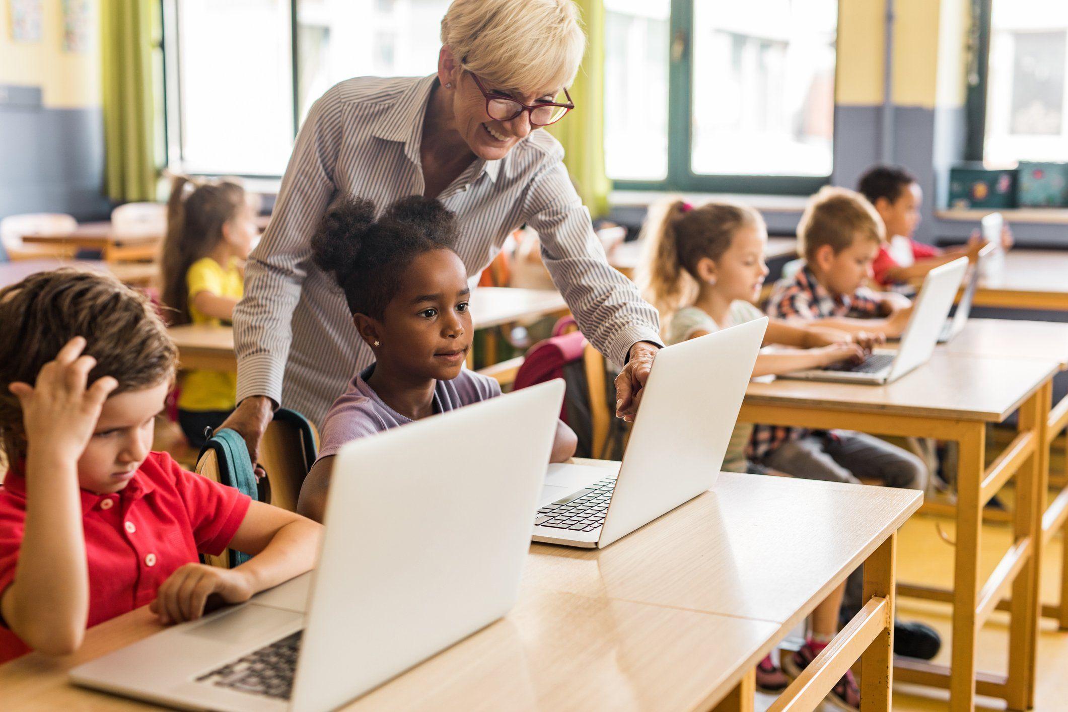 Classroom setting with a teacher standing up and pointing to a screen that is in front of a child sitting in their seat