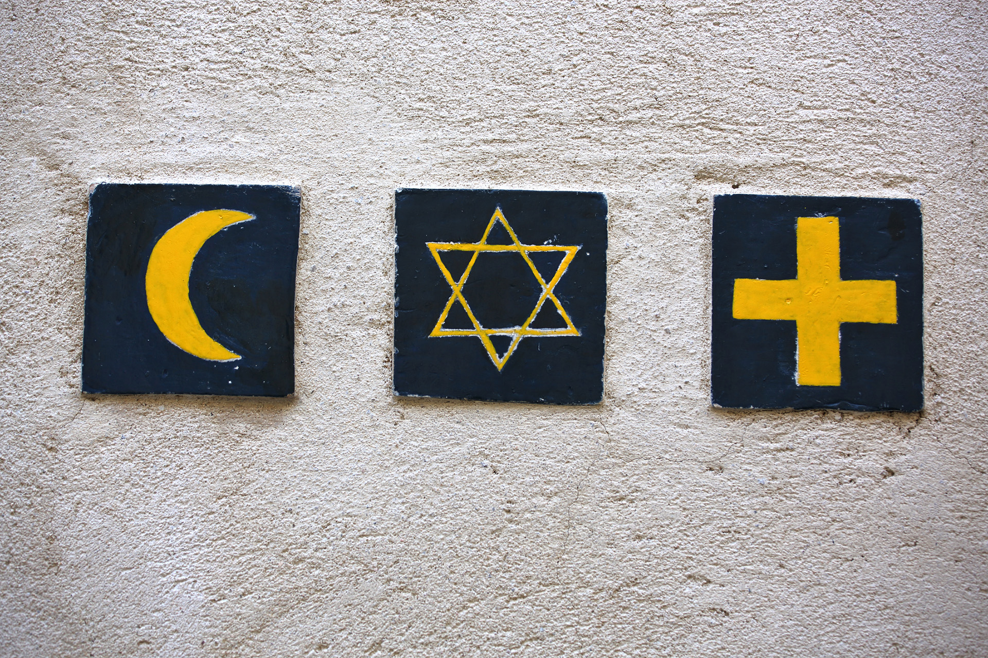 Three painted tiles showing a crescent moon, Star of David and cross on a textured wall.