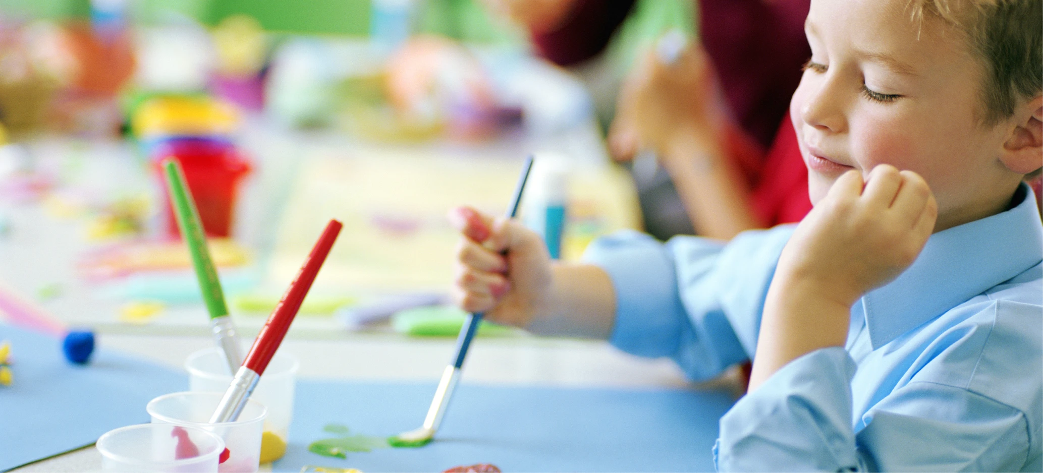 Boy in blue shirt paints with brushes at a classroom table, focused on his artwork with pots of paint nearby.