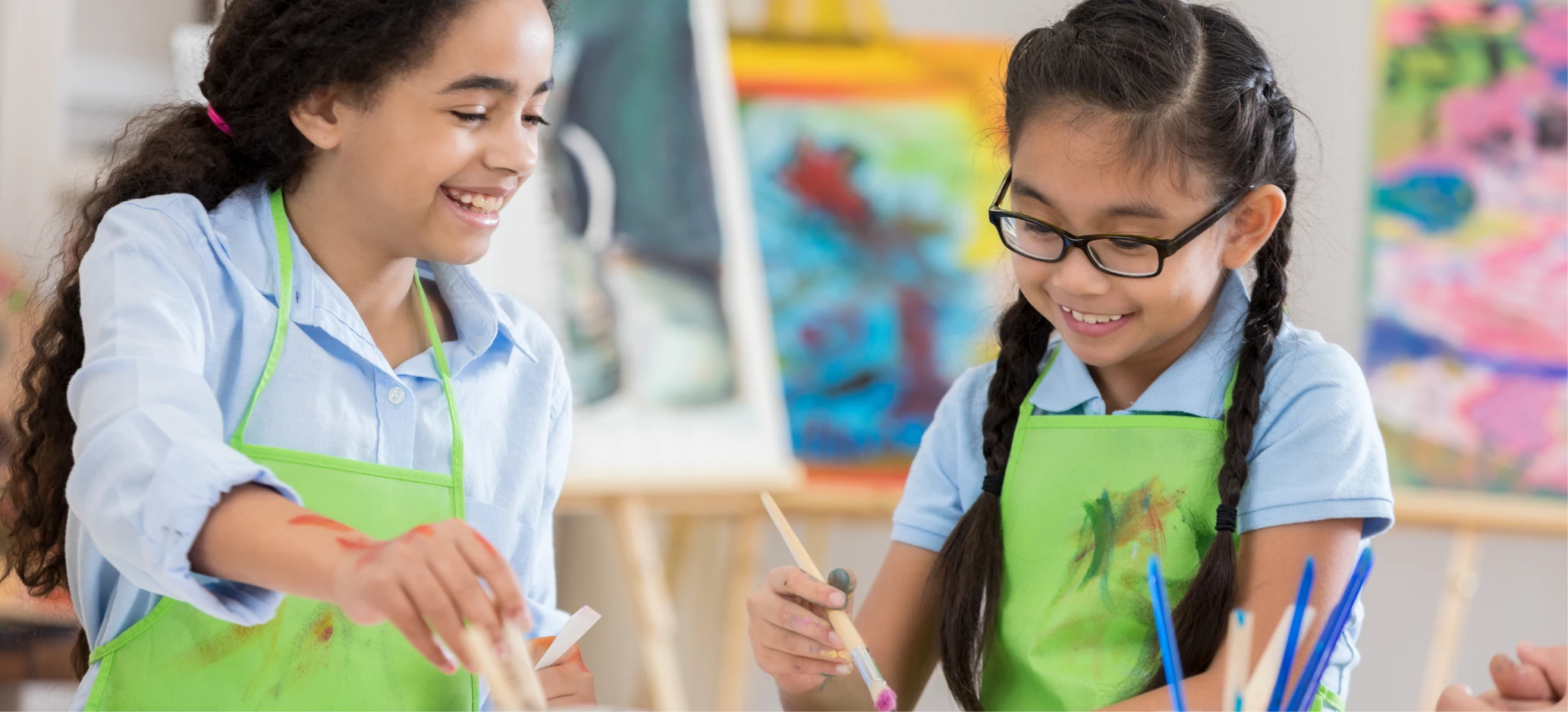 Two children in green aprons smile while painting with brushes during an art lesson, with colourful artwork in the background.