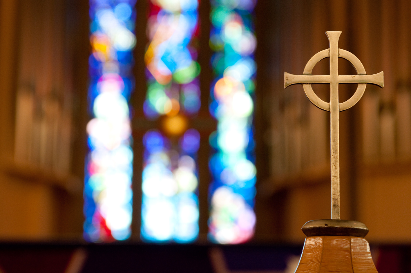 A cross with a blurred stained glass window in the background, creating a peaceful, spiritual atmosphere.