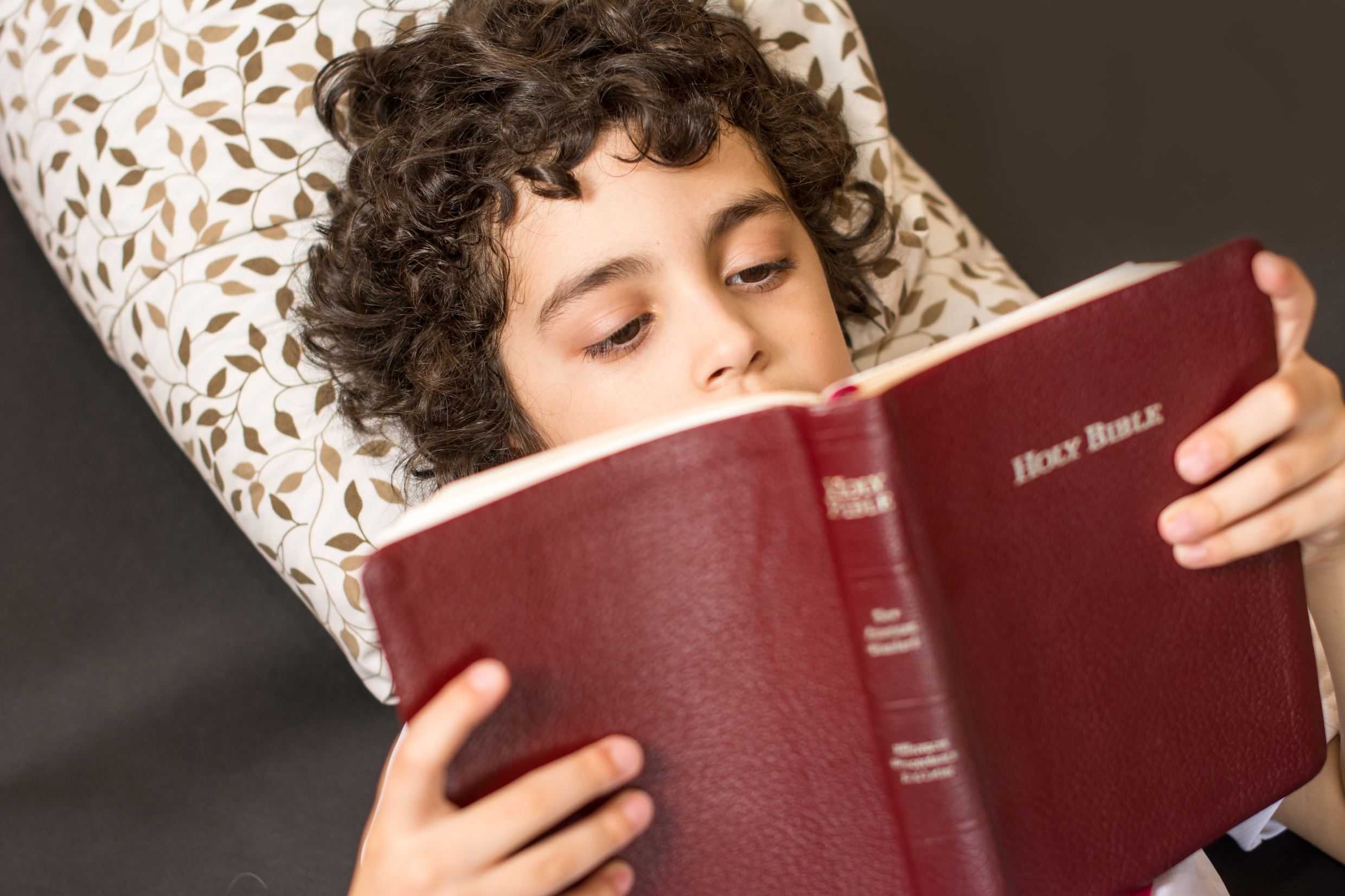 Child reading a red-covered Bible while lying on a pillow.