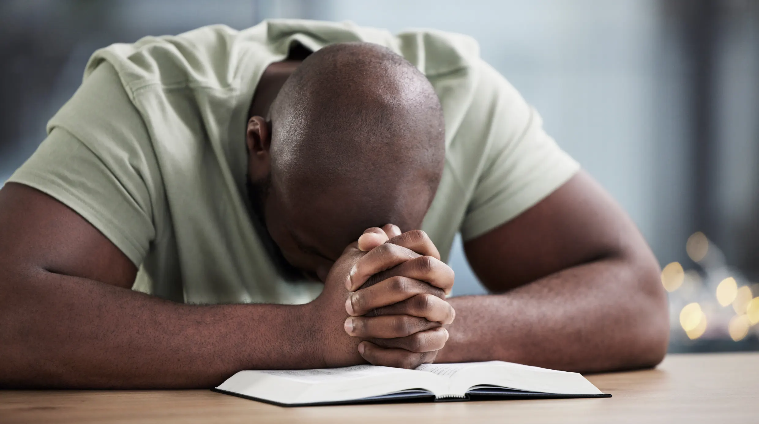 A man with his head bowed, resting on a book, with hands clasped together.
