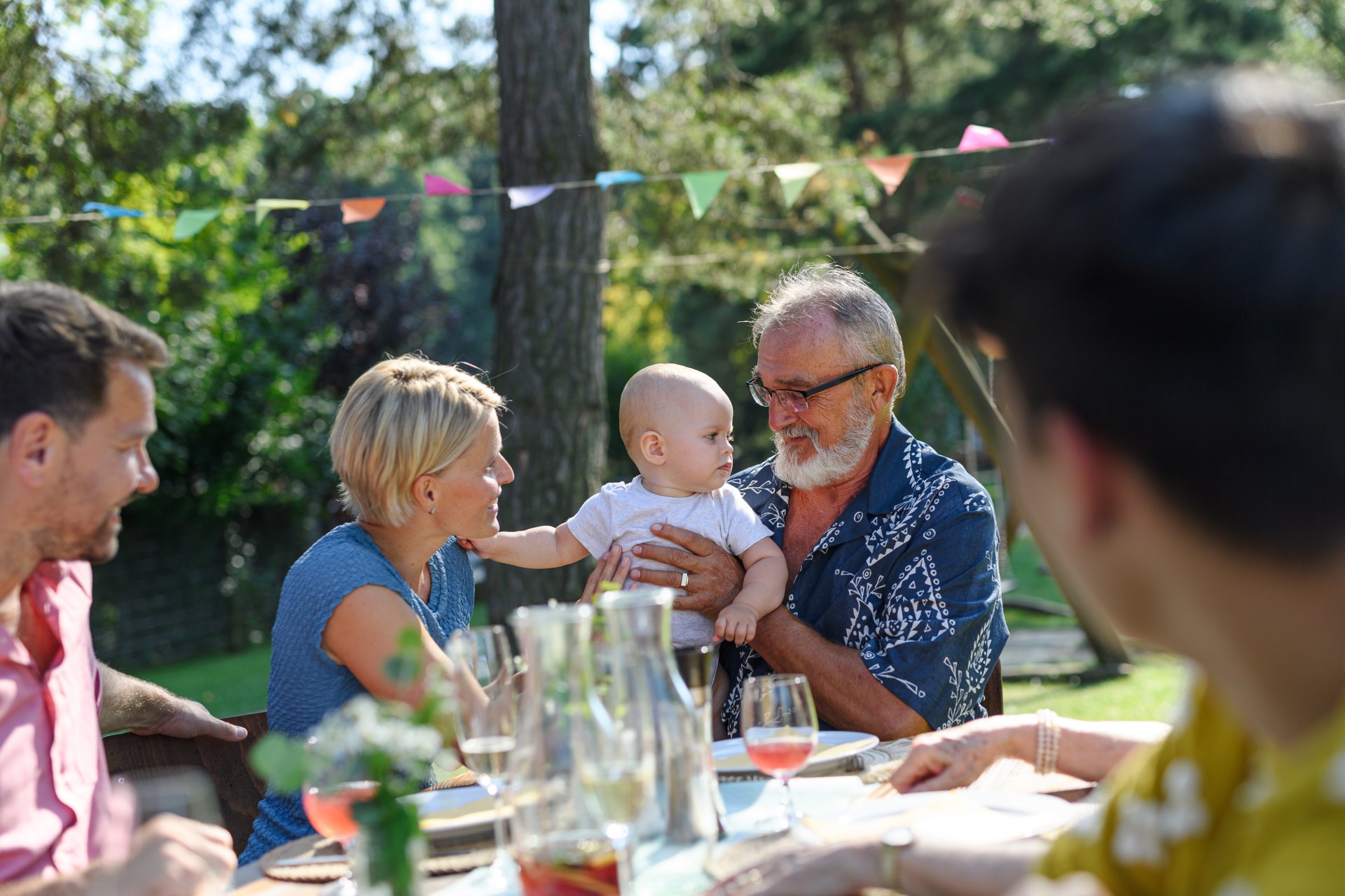 A man passes a baby to a woman. They are sat around a table in a garden.