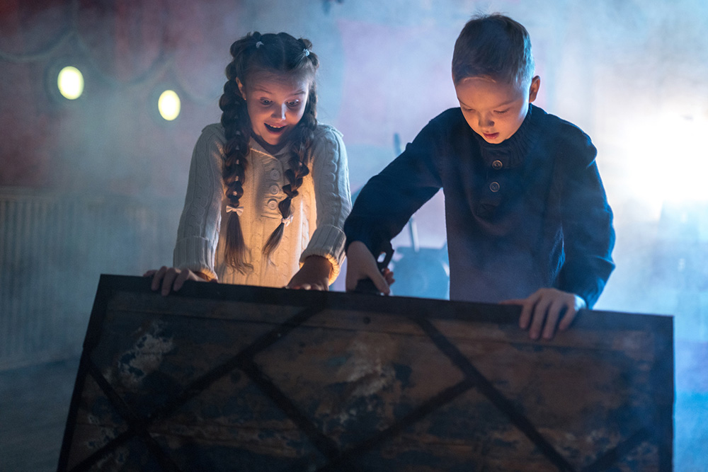 Two children look excited as they open a large wooden chest in a dark, smoky room.