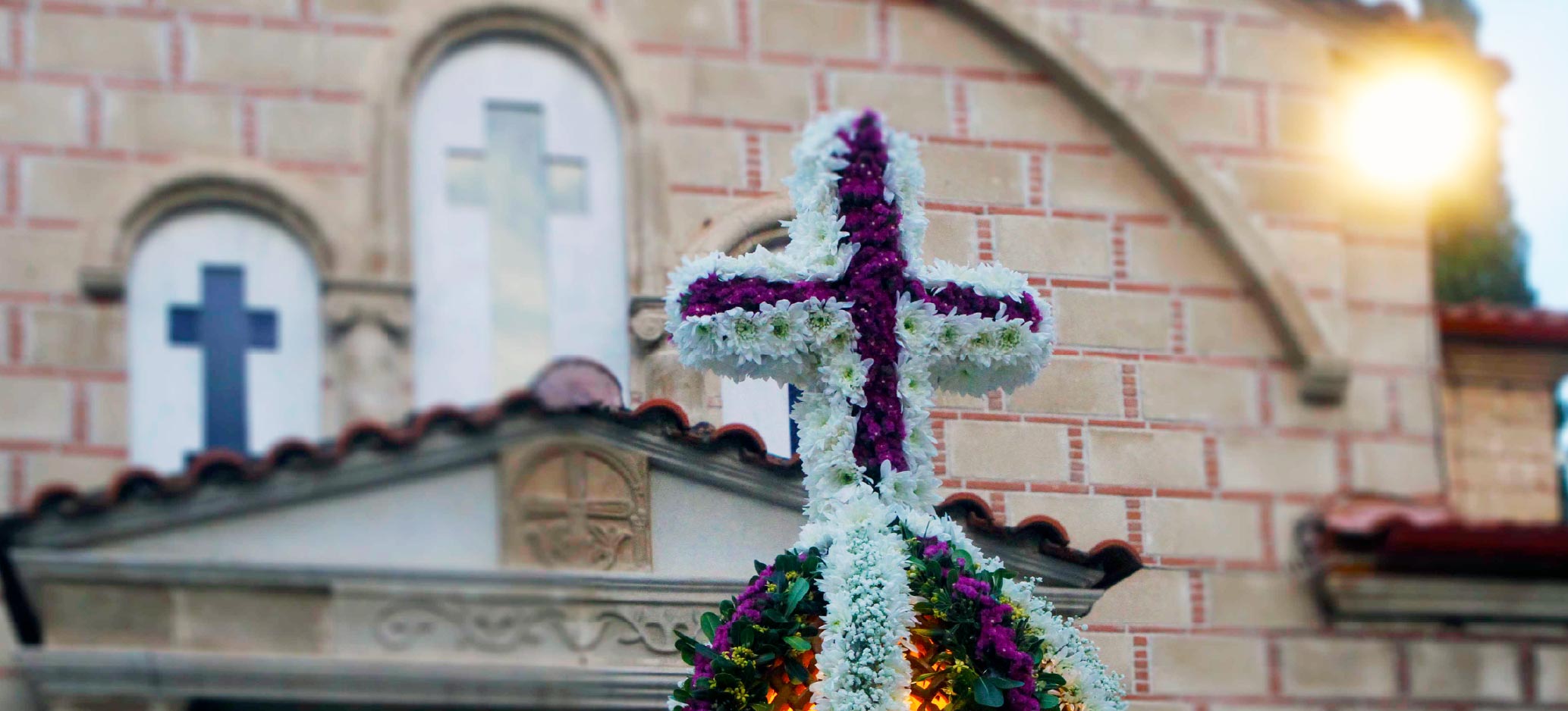 Floral cross decorated with white and purple flowers standing in front of a church building with cross-shaped windows.