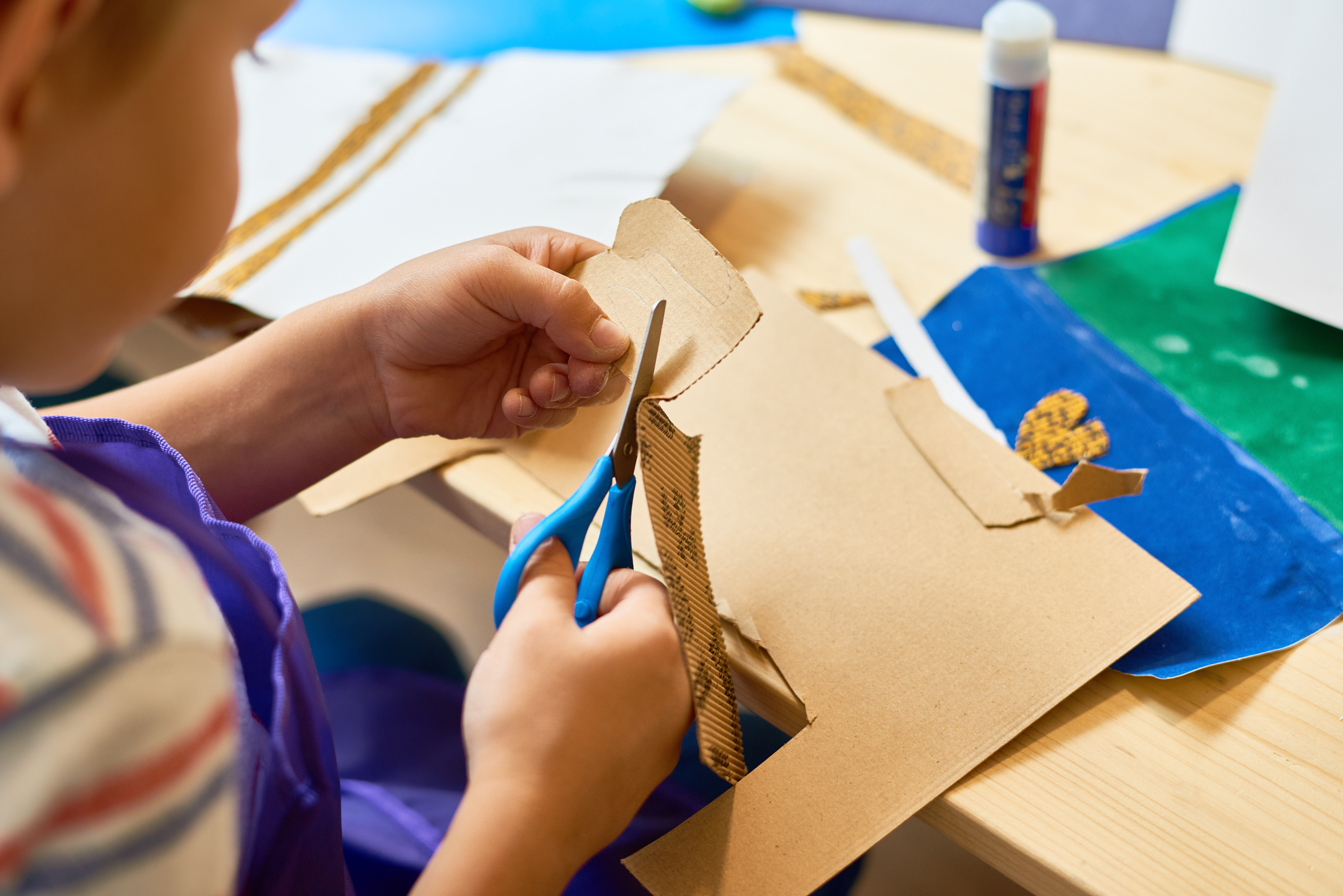 Child cutting brown paper with scissors and glue stick in the background.