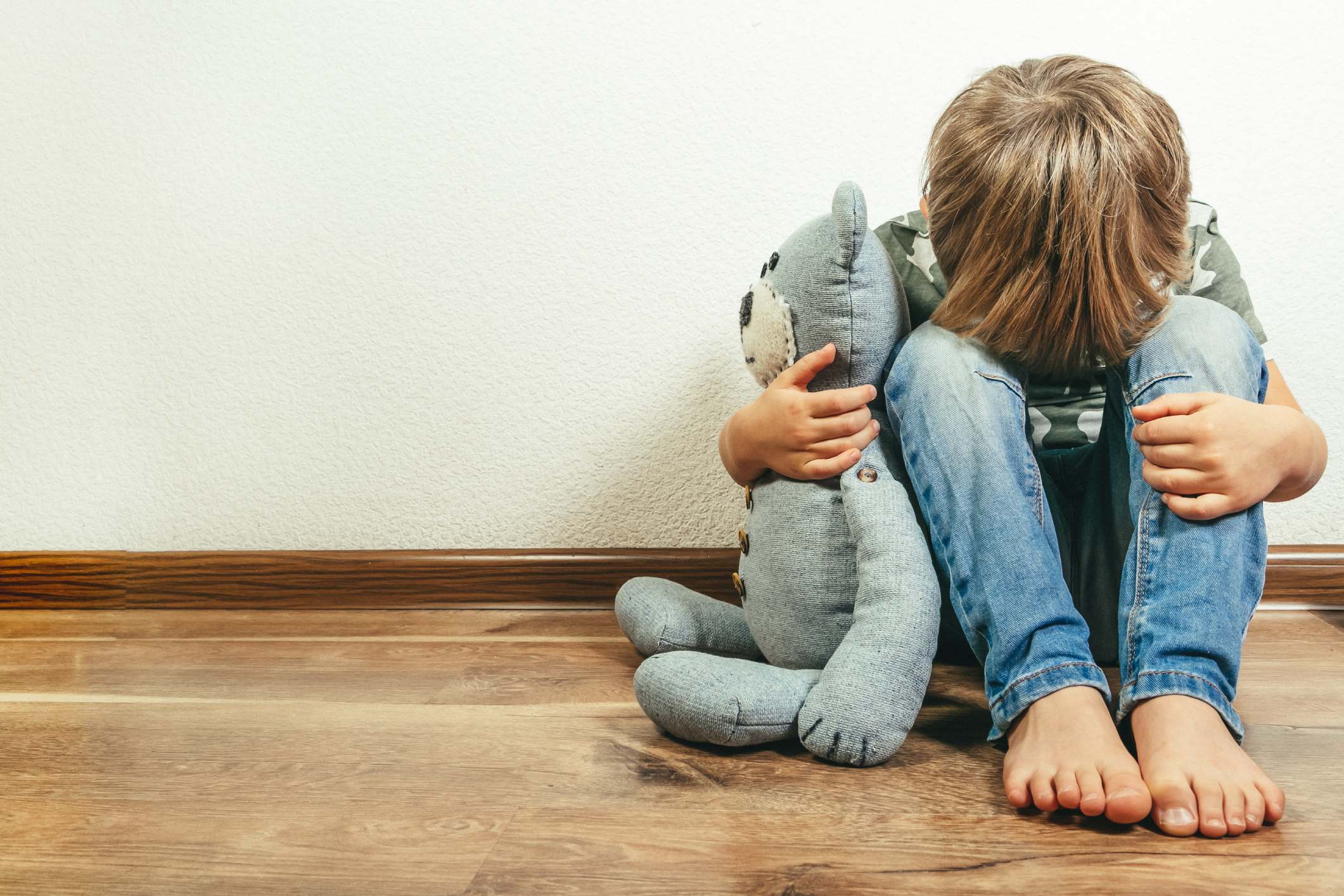 Boy sitting on floor hugging teddy bear looking upset
