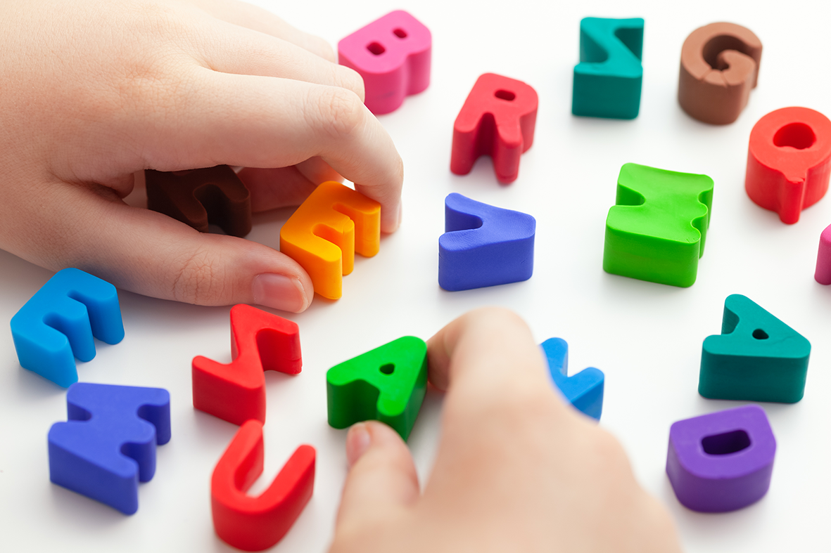 A child's hands pick up the letters E and A from a white desk of scattered plastic letters.