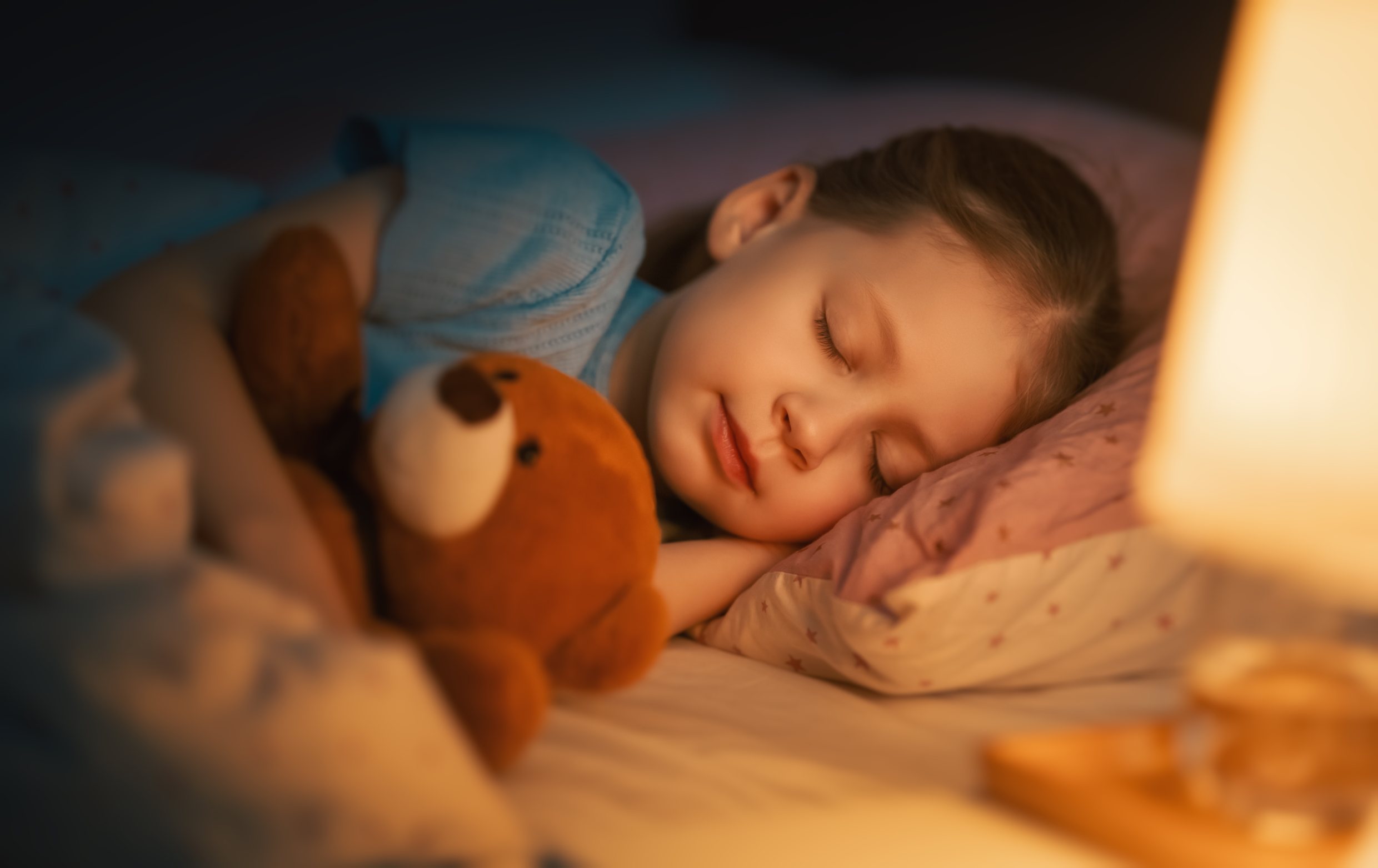 A young girl peacefully sleeping in bed while cuddling a brown teddy bear under soft lighting.