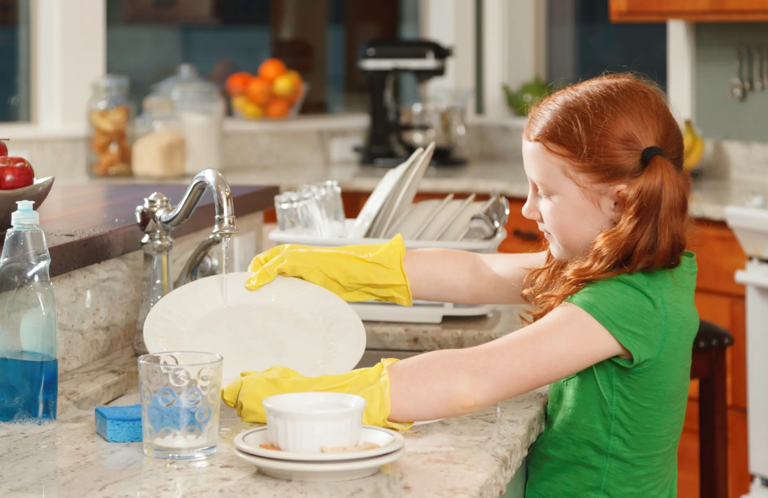 Child wearing yellow gloves washing dishes at a kitchen sink, helping with household chores.