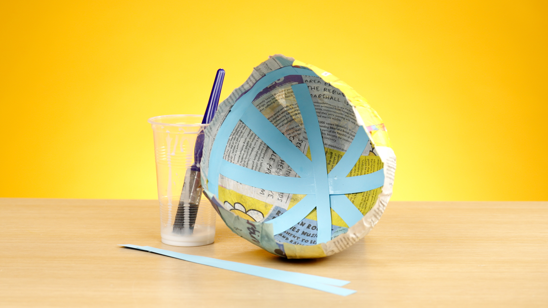 A photo of a papier-mâché structure, with a pot of glue and a brush next to it, against a yellow background.