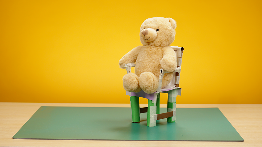 Light brown teddy bear sat on a chair made from cardboard tubes for legs and a cardboard seat. The chair has arms and it on a green mat against a yellow background.