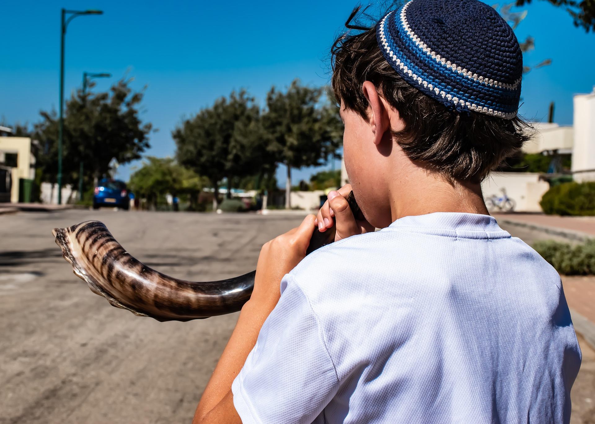 Boy wearing a kippah blowing a shofar outdoors on a sunny day.