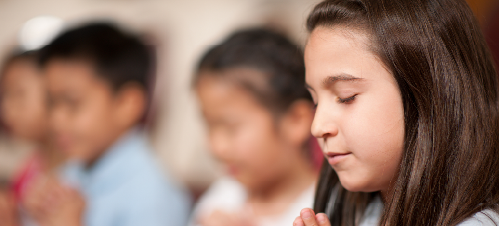 Child with eyes closed and hands together, appearing to pray, with other children blurred in the background.