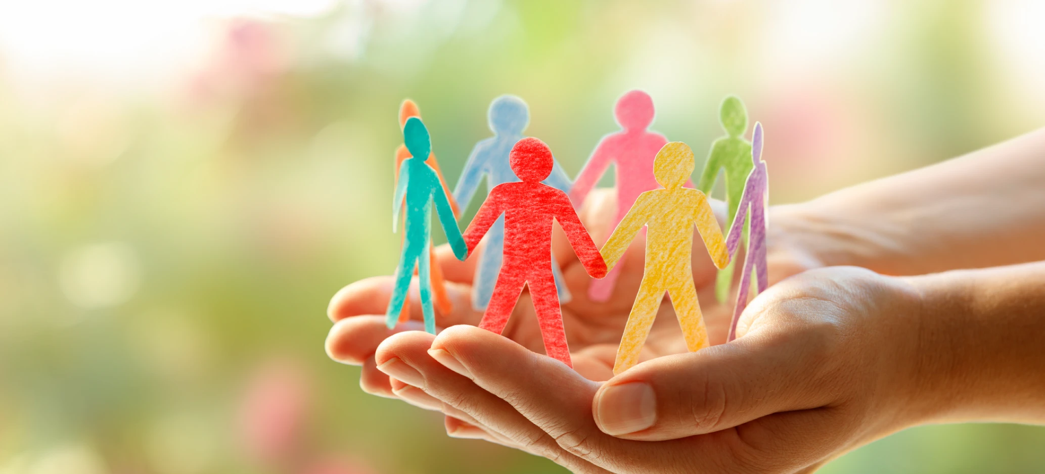 Two hands gently holding a row of colourful paper cut-out people linked together, with a soft, blurred outdoor background.