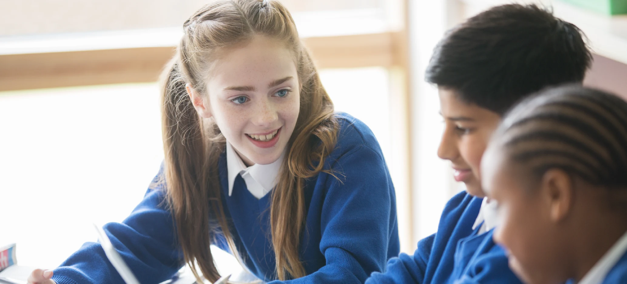 Three pupils in blue school uniforms smiling and working together at a desk.