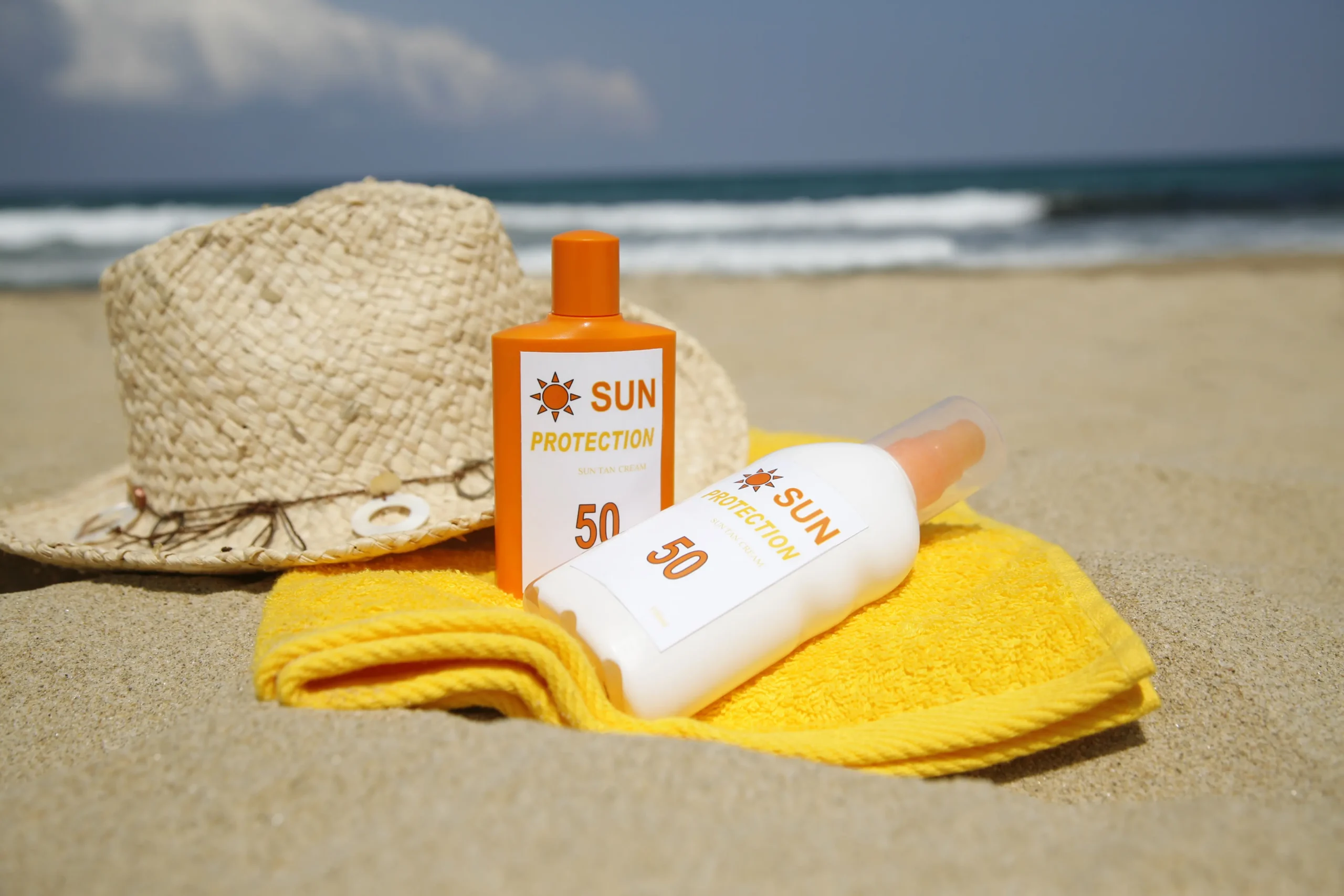 SPF 50 sun cream, a sun hat and yellow towel arranged on a sandy beach