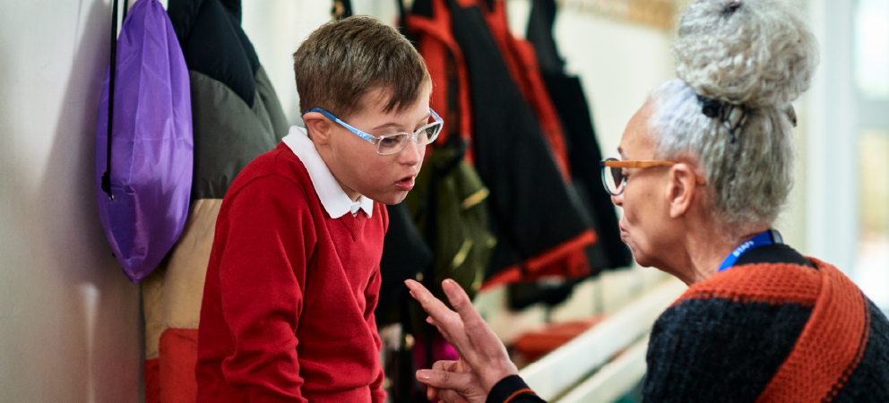 Photograph of a child talking to a teacher in the corridor of a school, wearing school uniform.