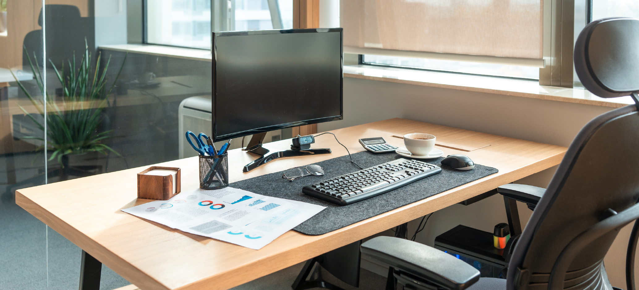 A modern office desk with a computer monitor, keyboard, mouse and webcam. The desk also contains a calculator, coffee cup, glasses, pens and printed charts, with an office chair beside it and large windows in the background.