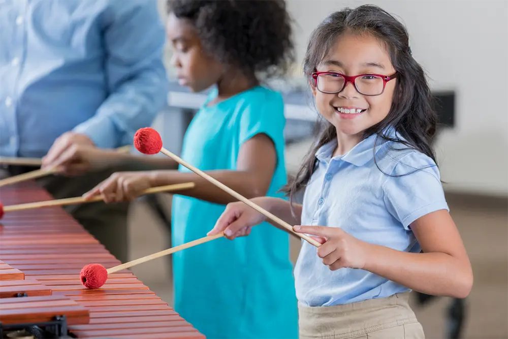 Two children play a xylophone with red-headed mallets during a music lesson while an adult supports the activity.