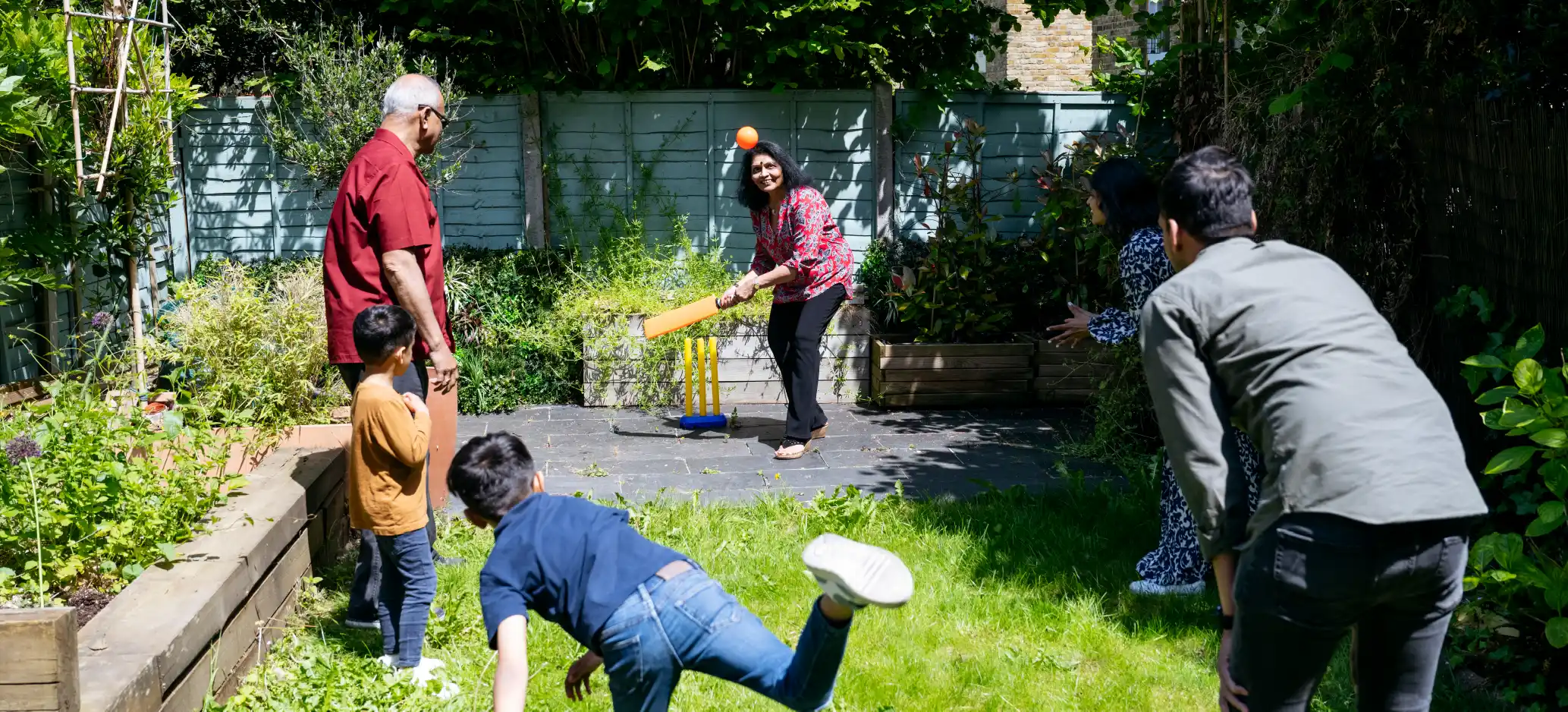 A family enjoys a game of garden cricket together, with adults and children taking turns batting, bowling and fielding in the sunshine.