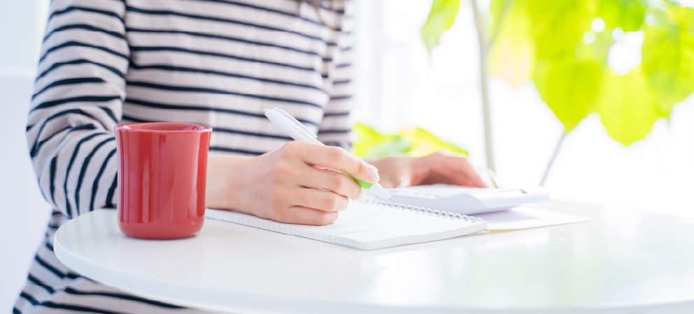 A person writing in a notebook at a table with a calculator and a red mug.