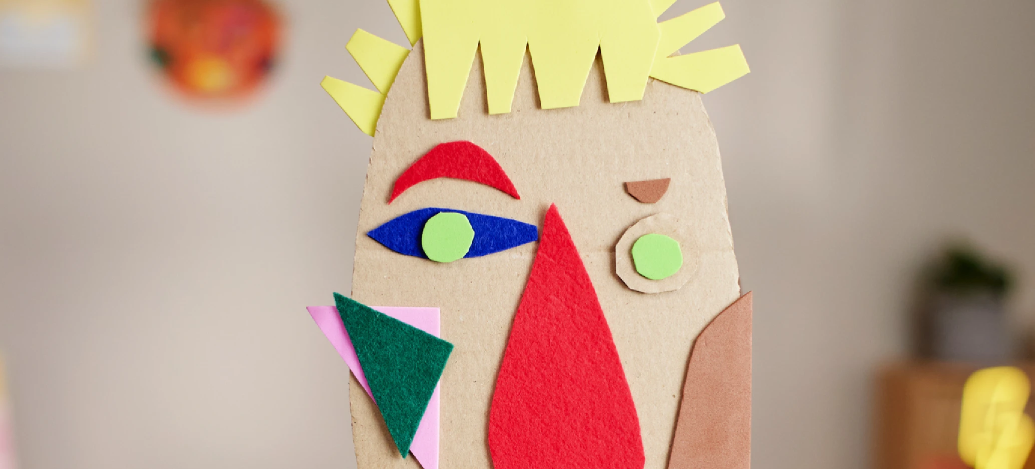 Close-up of a handmade cardboard face mask with yellow paper hair, mismatched eyes and colourful geometric felt shapes.
