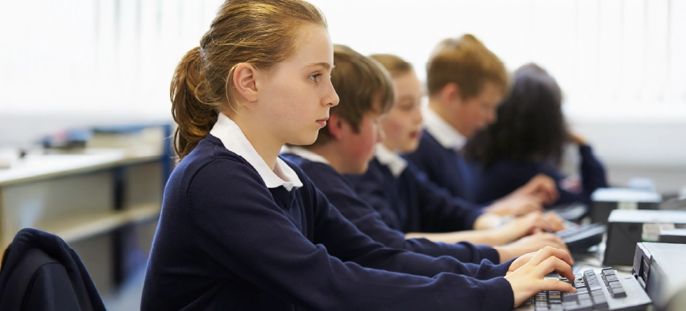 Pupils in school uniform use desktop computers in a classroom.