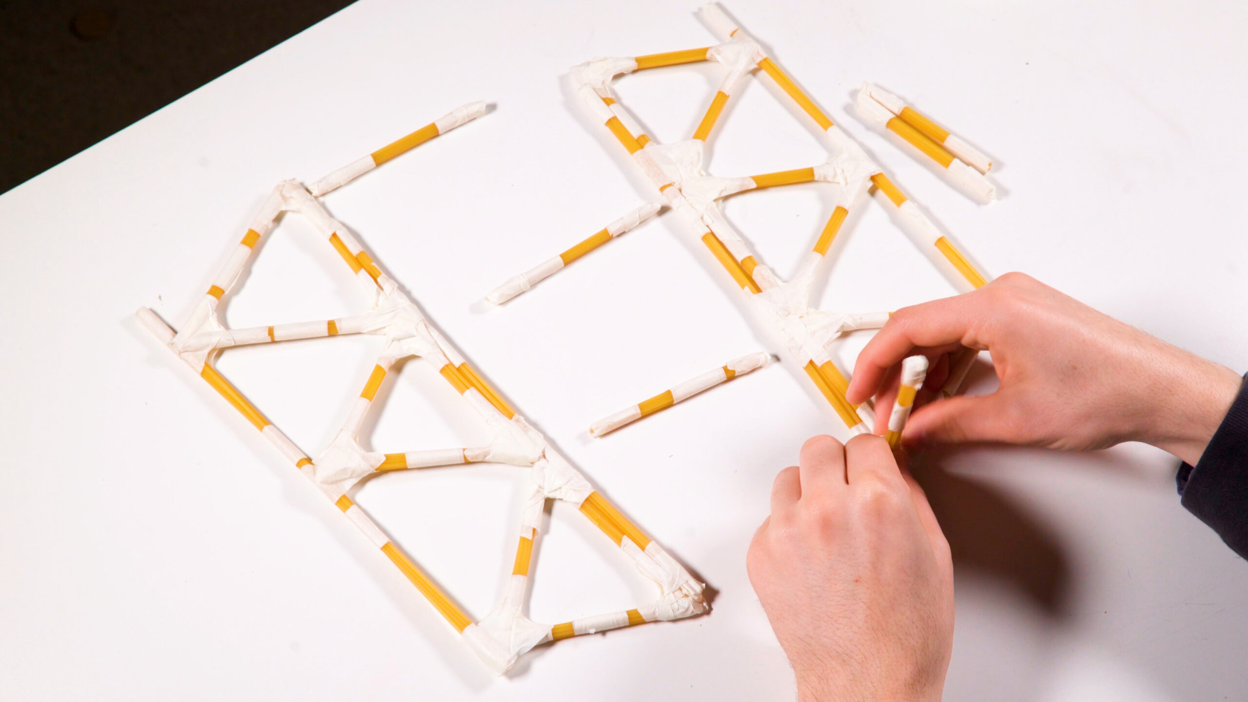 Hands assembling a small bridge model made from rolled paper tubes taped into triangles.
