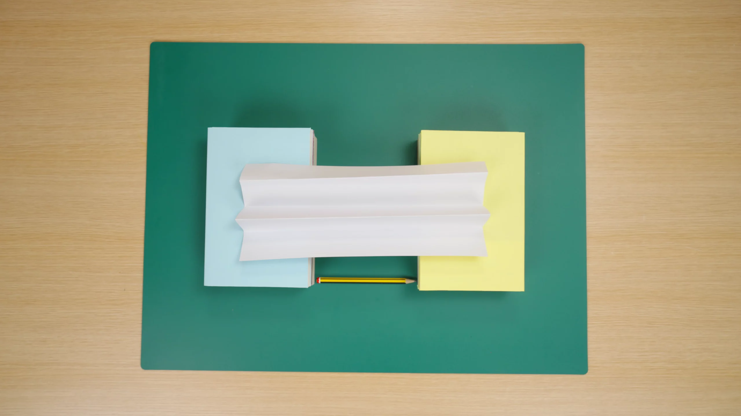 Two piles of books of equal height with a piece of A4 paper folded into a zigzag and places between the books like a bridge. There is a pencil between the books to show the distance.