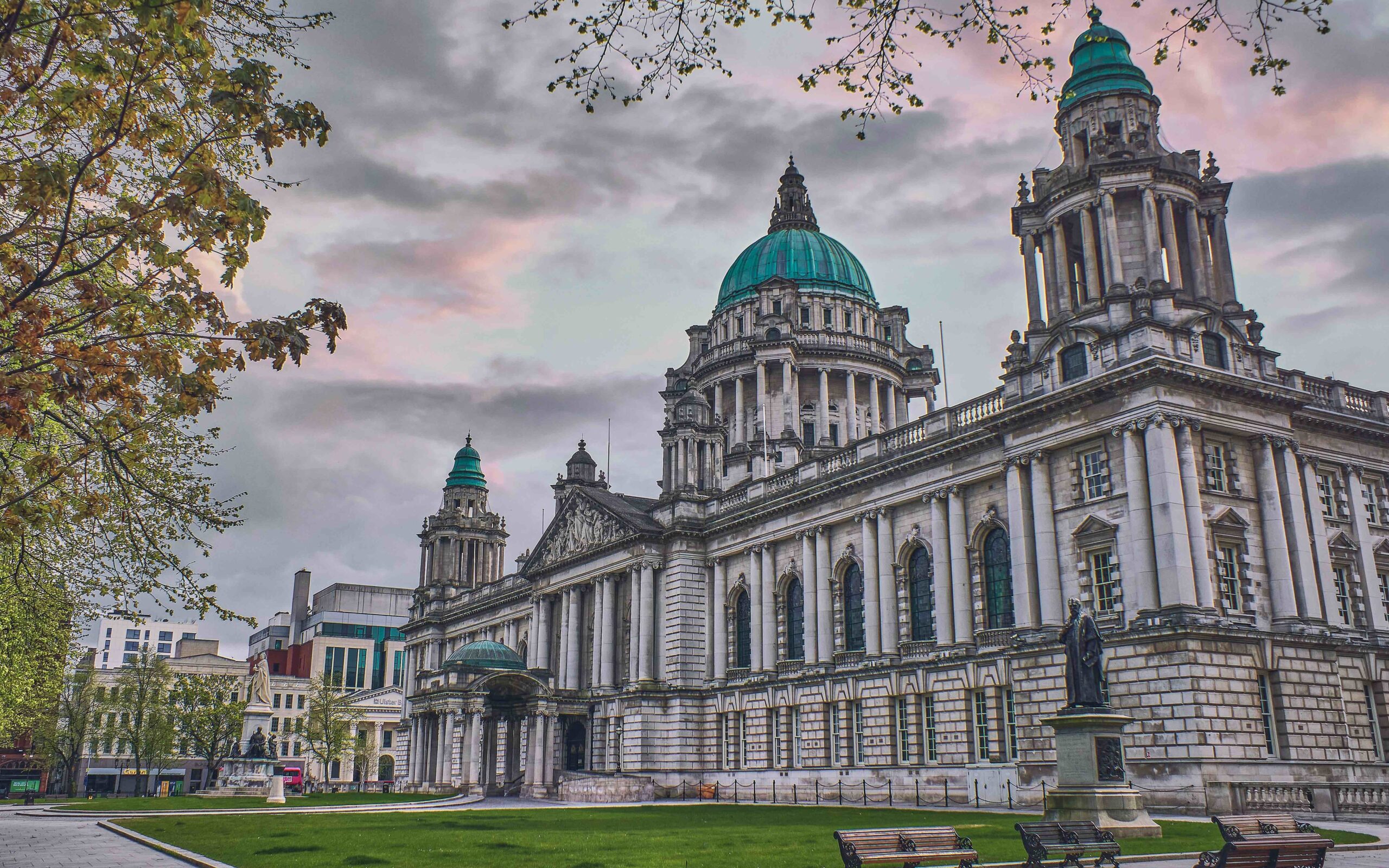 A large historic building with domes and columns surrounded by green lawns and trees under a cloudy sky.