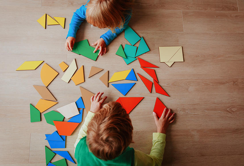 Two children playing with colourful tangram puzzle pieces on the floor.
