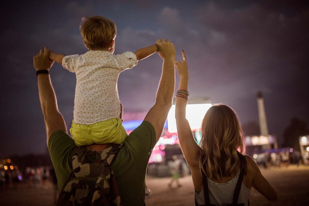 Parents and child enjoying a festival, with the child on the parent's shoulders.