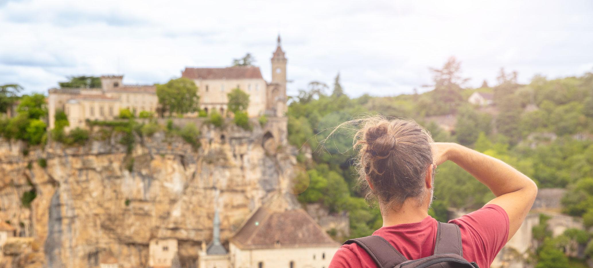 A man wearing a backpack looks out over a landscape of historic buildings, cliffs and forests.