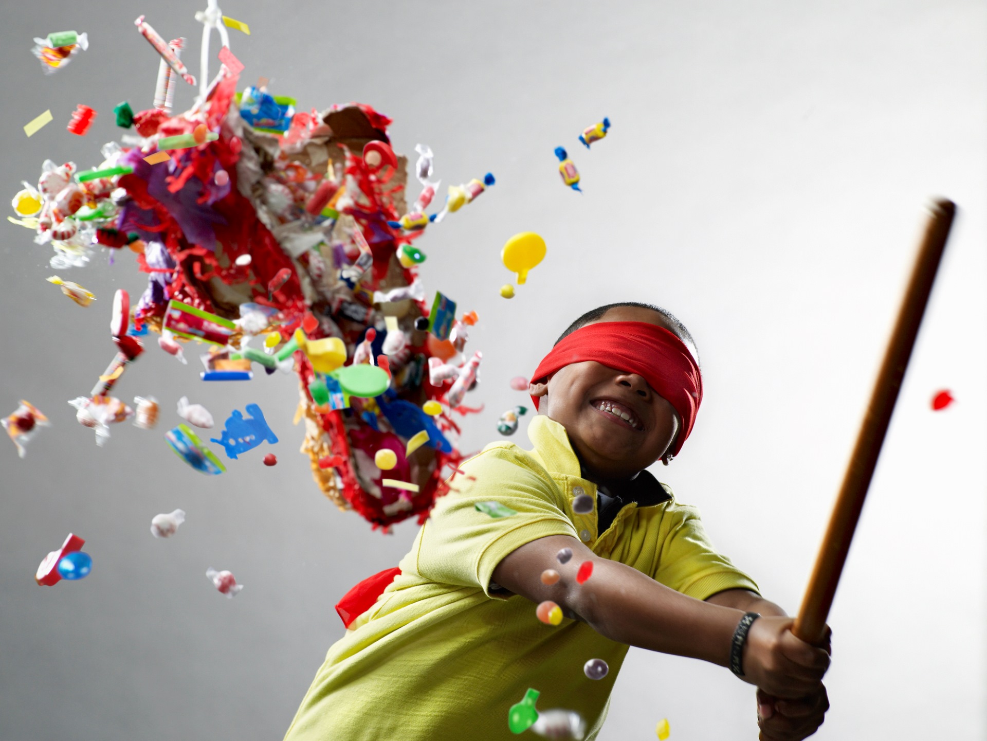 Smiling blindfolded child breaking colourful piñata with sweets and toys flying