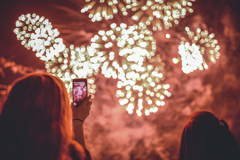 People watch fireworks in the night sky, with one person holding up a phone.