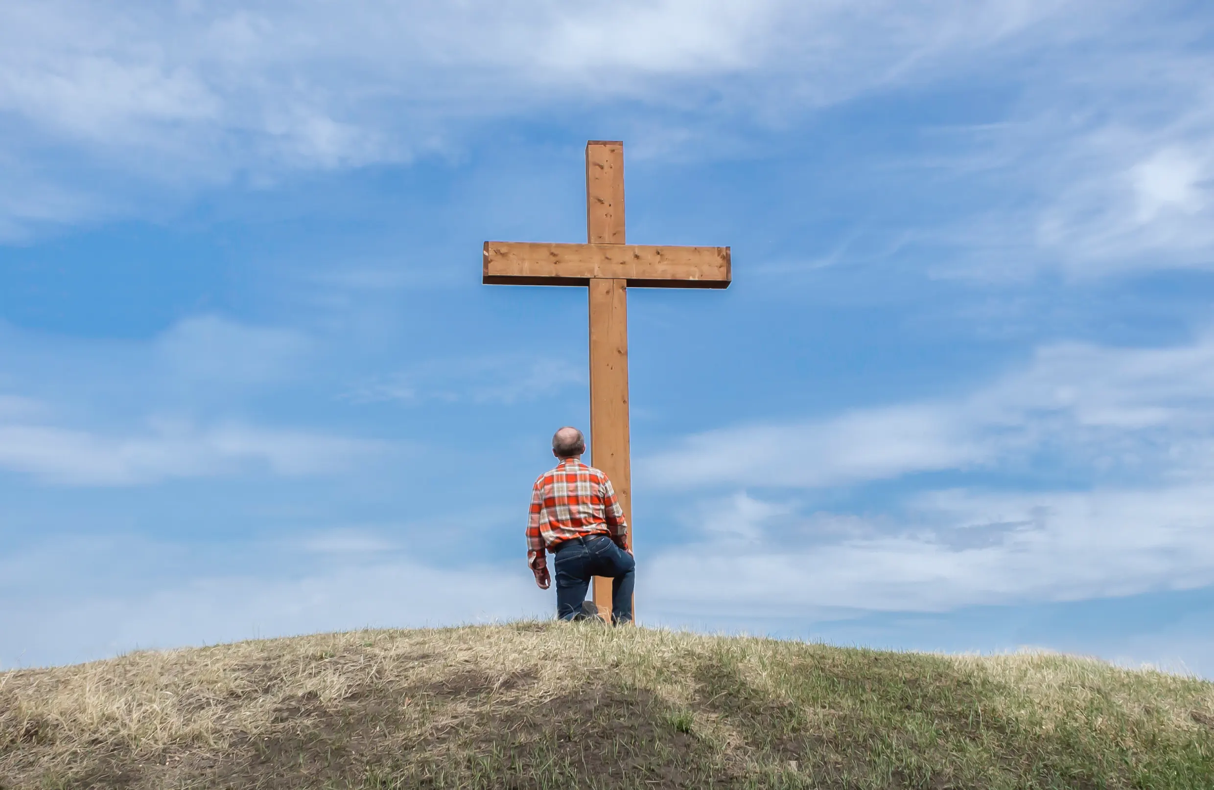 A man stands at the foot of a large wooden cross on a hill, looking up at it.