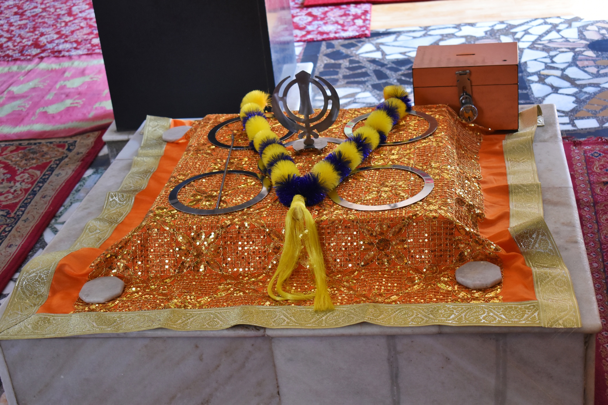Sikh religious display featuring the Khanda symbol, ceremonial items and a collection box on a decorated platform.