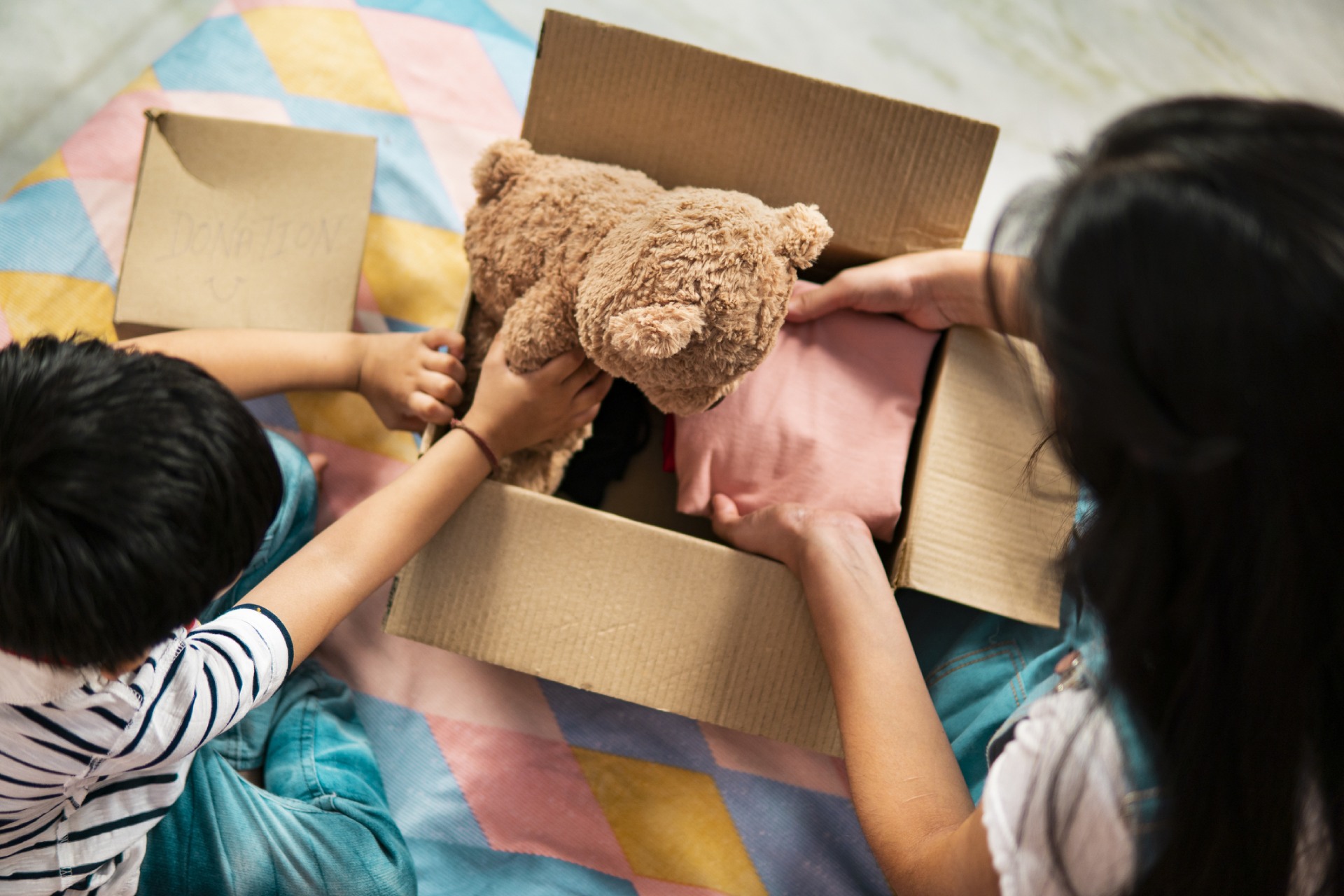 Two people sit on a bed and put a teddy bear and an item of clothing into a cardboard box.