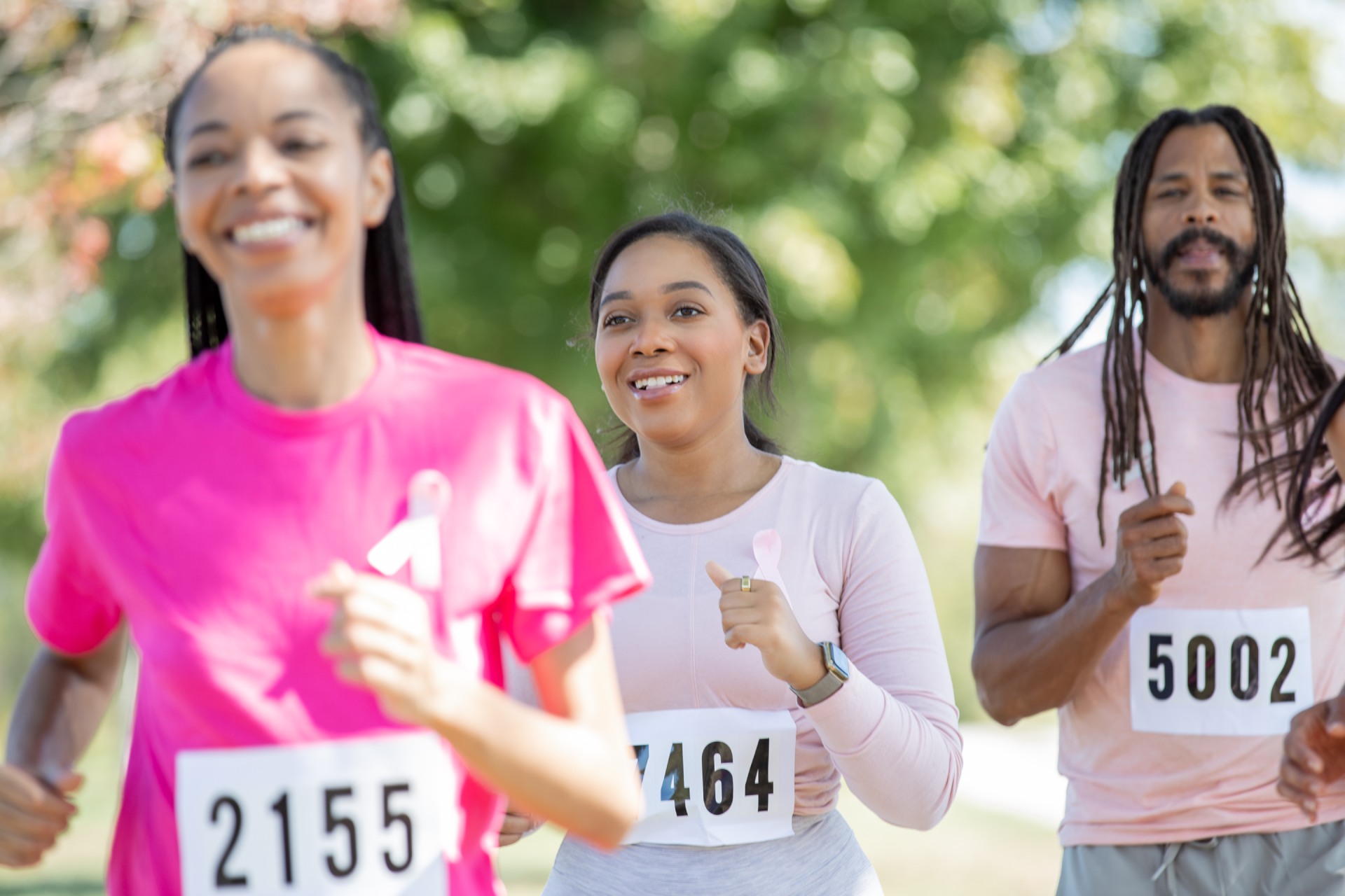 Three happy people wearing pink tops run in a charity race. They have numbers attached to their tops.