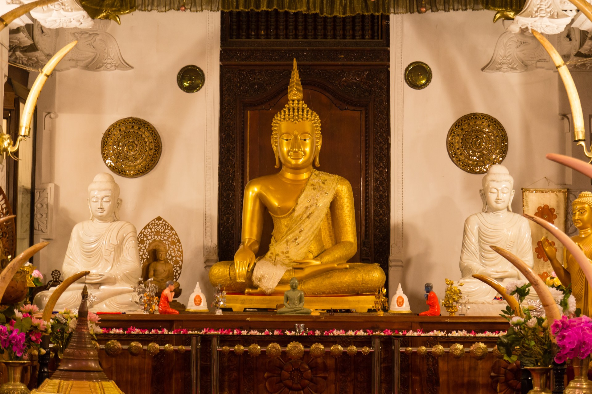 A large golden Buddha statue sits between two smaller white Buddha statues on a platform surrounded by tusks and flowers.