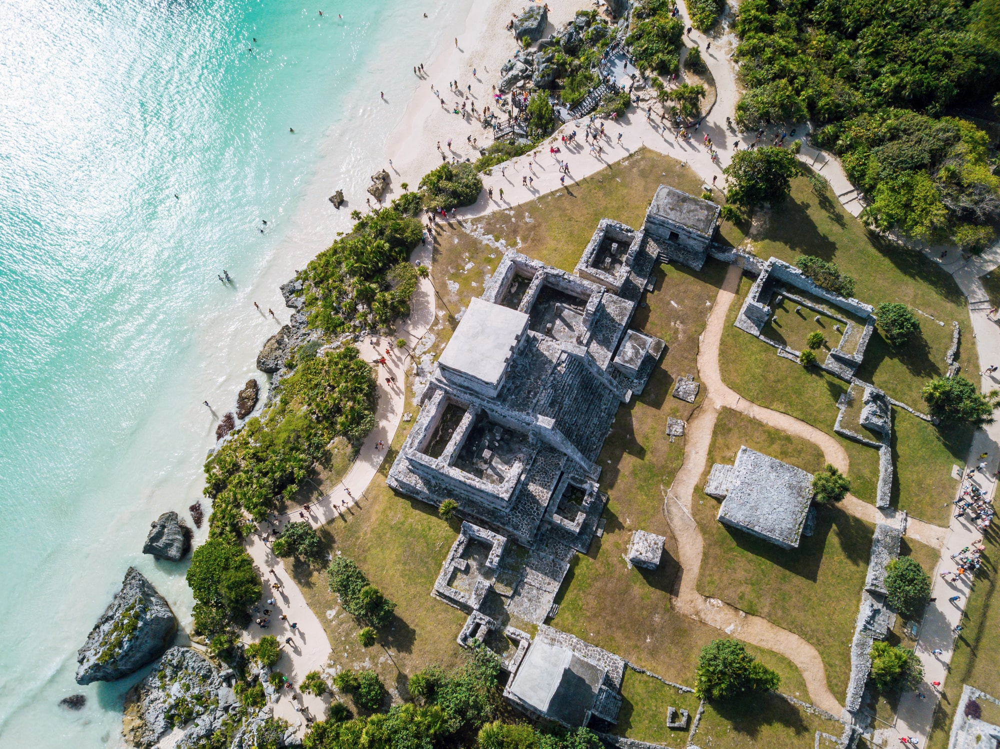 An aerial view of a group of Mayan buildings, with a beach and ocean to the left of the buildings. There are trees and plants around the beach and buildings.