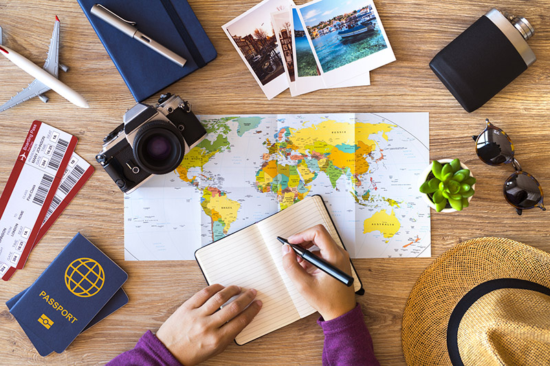 A person writing in a notebook beside a world map and travel items on a table.