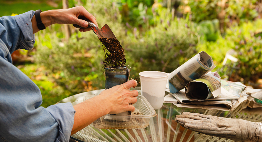 A person uses a trowel to fill a pot with soil. They are sitting at a table covered in gardening tools. They are in a garden.