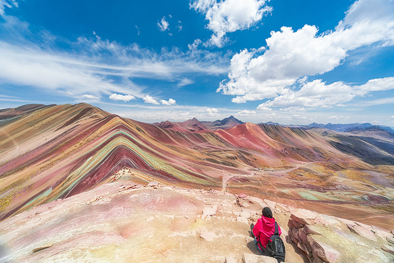 A mountainous landscape with brightly coloured striped hills under a blue sky, with a person sitting in the foreground.