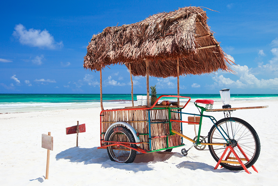 A smoothie bar attached to a bike is on a tropical beach, with a clear blue sky