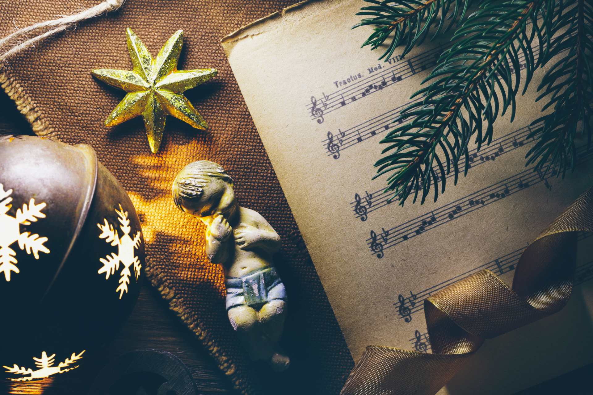 A figurine of the baby Jesus lies on a piece of fabric, as well as a sheet of music, a branch of a pine tree, golden star, some ribbon and a bauble